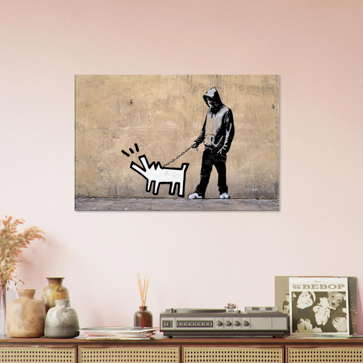 Banksy Choose Your Weapon and Keith Haring barking dog canvas displayed above a sofa in a warm peach-toned living room.