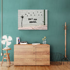 Banksy Don't Stop Dreaming graffiti canvas displayed above a cabinet against a teal wall.
