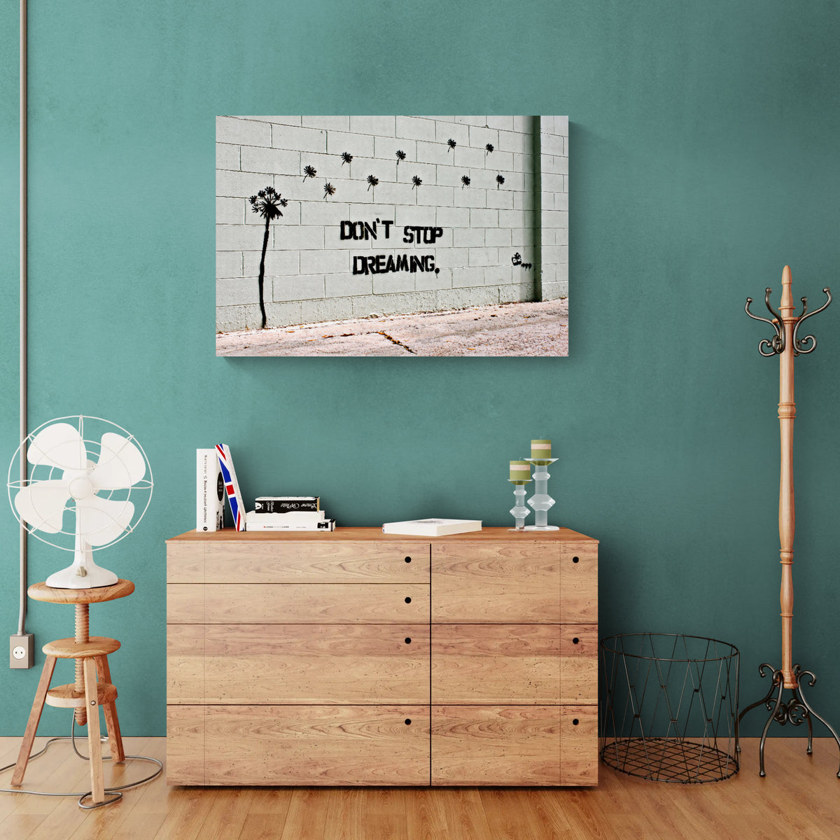 Banksy Don't Stop Dreaming graffiti canvas displayed above a cabinet against a teal wall.