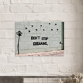 Banksy Don't Stop Dreaming canvas wall art displayed against a white brick wall.